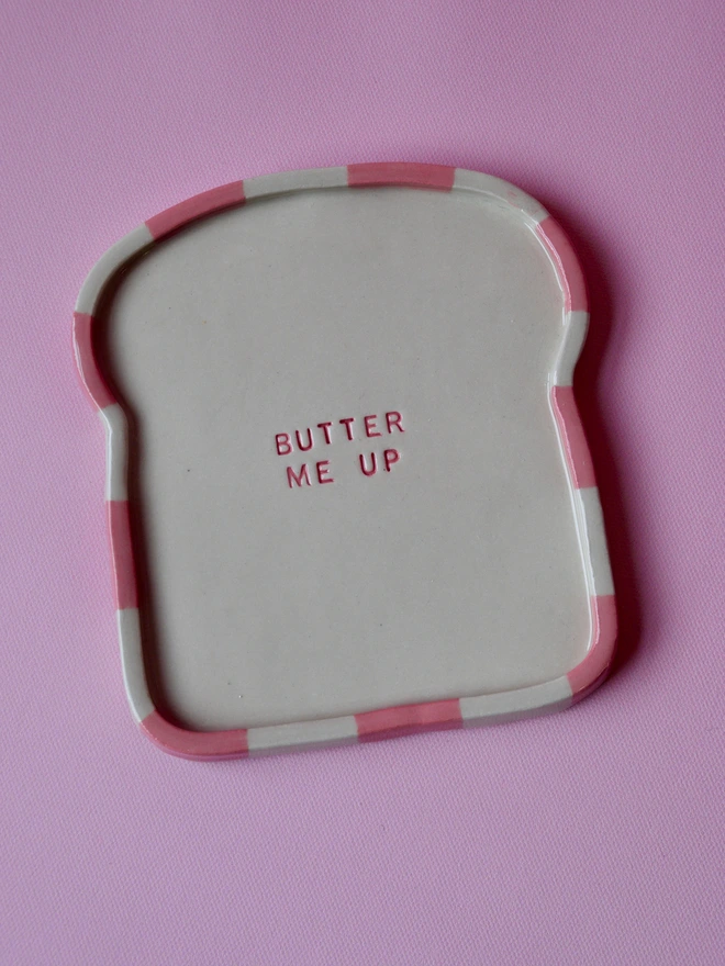 Handmade ceramic toast plate shaped like a slice of bread, featuring a pink striped rim and the words “Butter Me Up” stamped in the centre, photographed on a pink background.