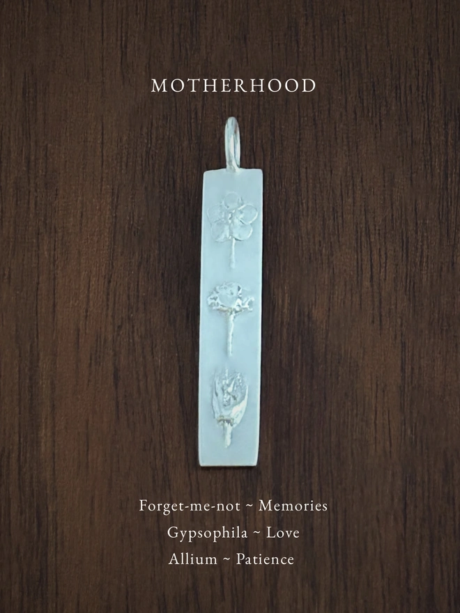 Motherhood bar pendant in sterling silver on a dark background with the meaning of the flowers written in white