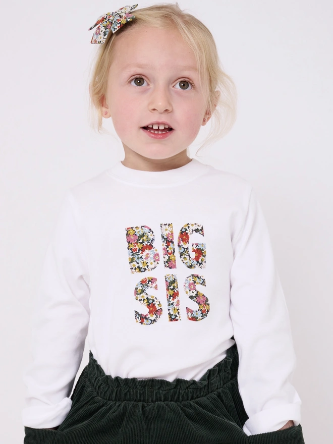 a little girl in a white t-shirt that says Big Sis in the front in floral print