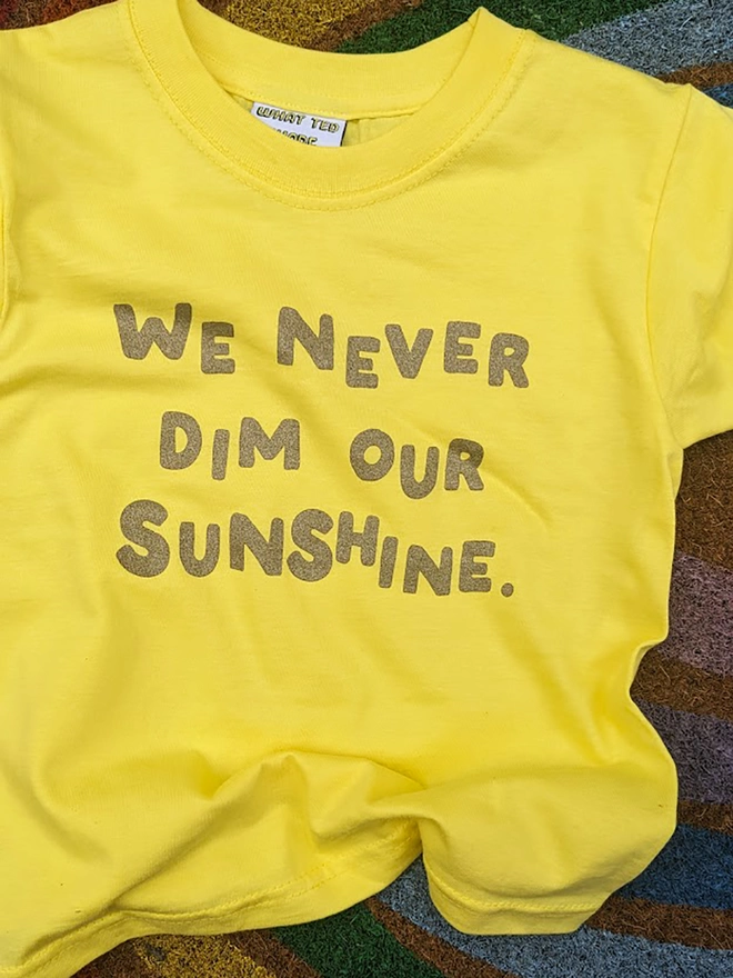 A yellow t-shirt with the slogan We Never Dim Our Sunshine  displayed on a rainbow mat