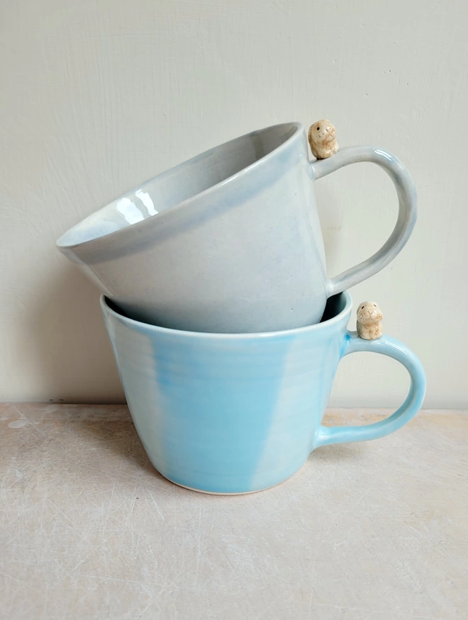 2 stacked pottery cups in pale blue and grey glaze with a miniature bunny on each handle