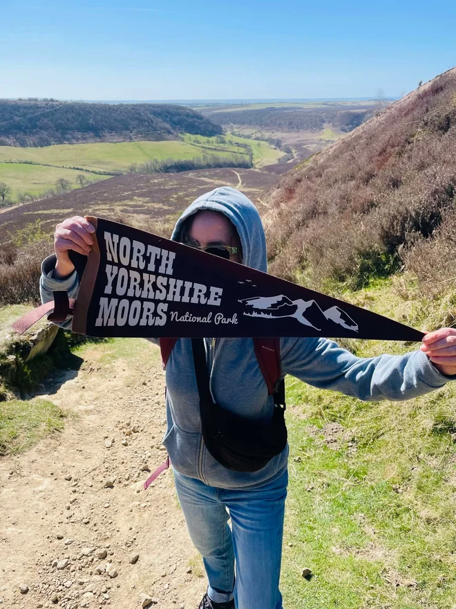 someone holding a flag pennant with the text North Yorkshire Moors, she is stood in the middle of the national park on the moor
