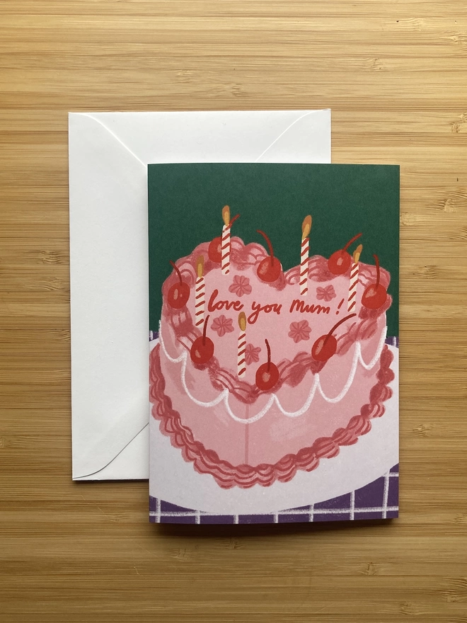 Greetings card on table, the design is a pink cake, with the words 'Love you Mum' iced on it