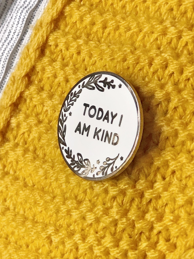 A round white enamel pin with a floral design and the words "Today I Am Kind" is pinned to a yellow cardigan.