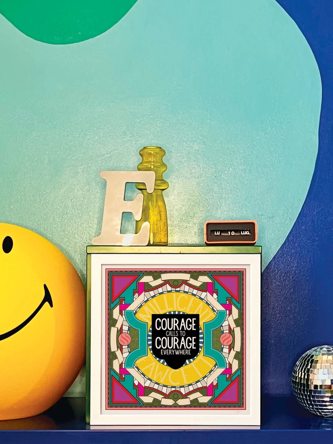 An illustration with “Courage calls to courage everywhere” written in white against a black background at the centre, surrounded by Millicent Fawcett written in yellow, and bordered with a symmetrical design in white, greens and pinks. It is framed in a white frame and is propped against a turquoise and dark blue wall. Next to the frame is a disco ball, a letter ‘E’ ornament, a yellow glass vase, an orange Italian plastic calendar showing the date as ‘LU 10 LUG’ and a large light up yellow Smiley lamp.