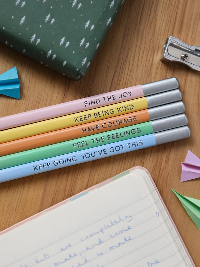 Five pastel coloured pencils placed on a wooden desk next to a silver pencil sharpener. Each pencil has a positive message stamped in silver block foiling.