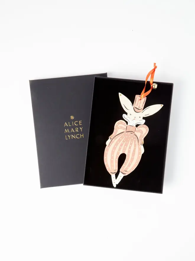 Hare Tree Dancer. wooden hanging hare decoration in an open black gift box embossed "alice mary lynch" in gold, with a pink top hat, dusky pink polka dot pantaloons with a bow and orange ribbon with a jingle bell