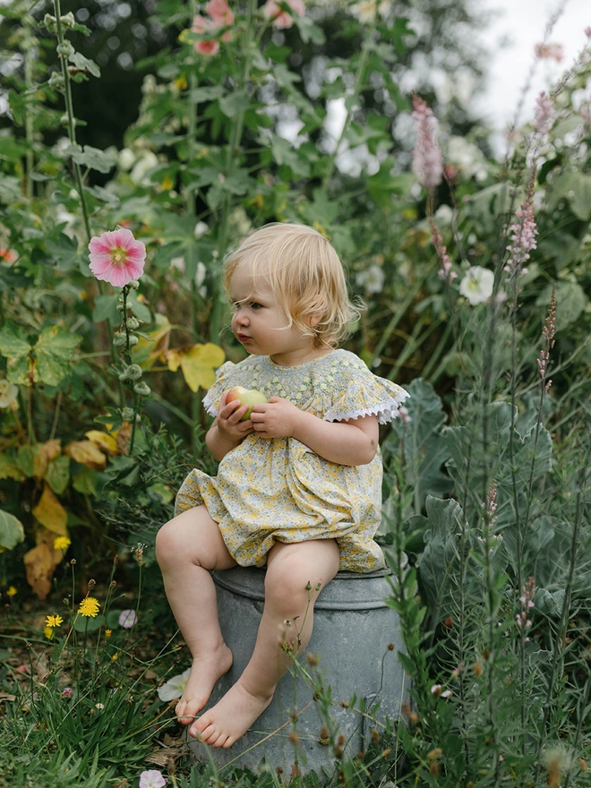  A toddler sits on an upturned metal bucket in a garden, surrounded by flowers and with an apple in hand wearing a yellow floral smocked romper.