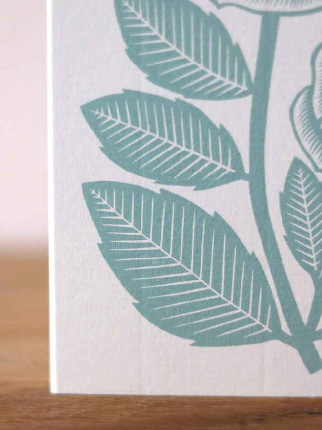A greetings card feature a teal rose design based on an original linocut print