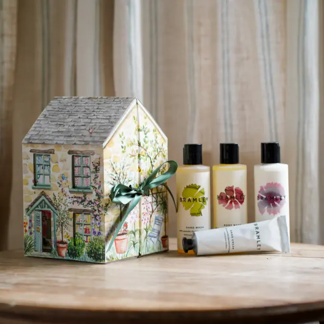 yellow cardboard cottage-shaped box with illustrated design and green bow, next to three bottles of product with white lables and black lids, and one white squeezetube