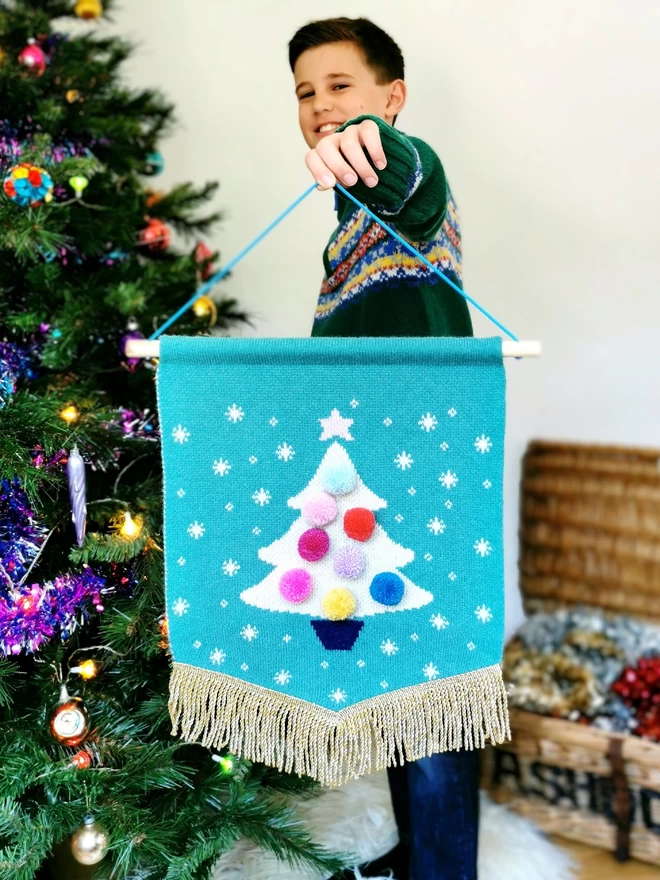 A boy wearing a colourful fairisle jumper stands next to a Christmas tree with retro coloured baubles. He is holding up a jade green knitted wall hanging with a white sparkly christmas tree knitted on the front. The tree is adorned with merry and bright rainbow pom poms and edged with a gold bullion trim. He is smiling and looking excited for the festive season.