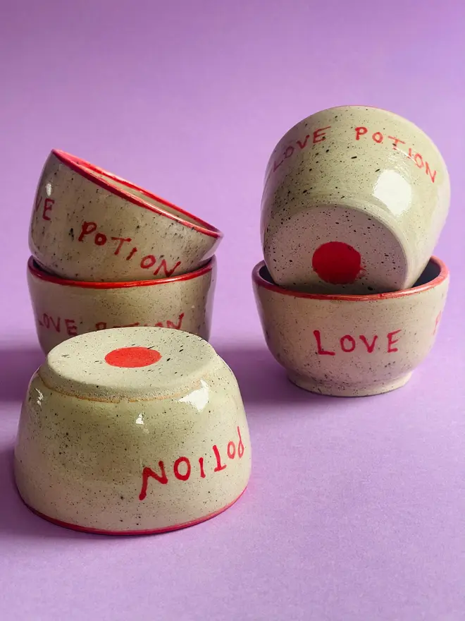 'Love Potion' Mug, a group of ceramic mugs that say 'Love Potion' in front of a purple background. Each mug is stamped with a signature red dot on the bottom. 