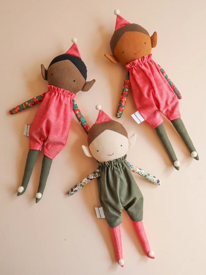 Three handmade Christmas elf dolls with pointed ears, wearing olive green romper suits with colourful floral Liberty print sleeves, red hats and matching long legs with white pom-pom feet. Light, mid and dark skin tones.