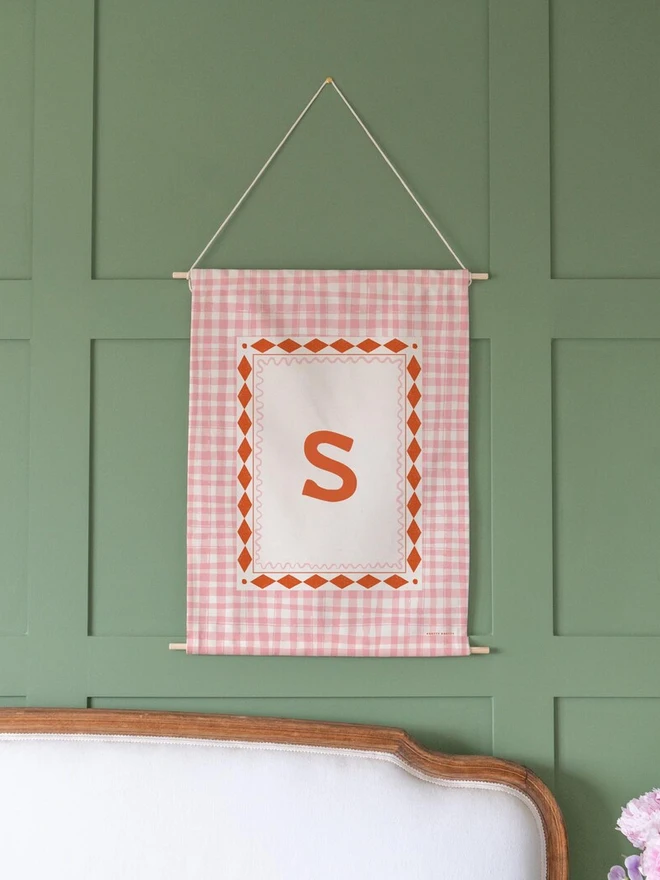 Personalised Gingham Initial Fabric Banner, in pink and red hanging on a green wall. 