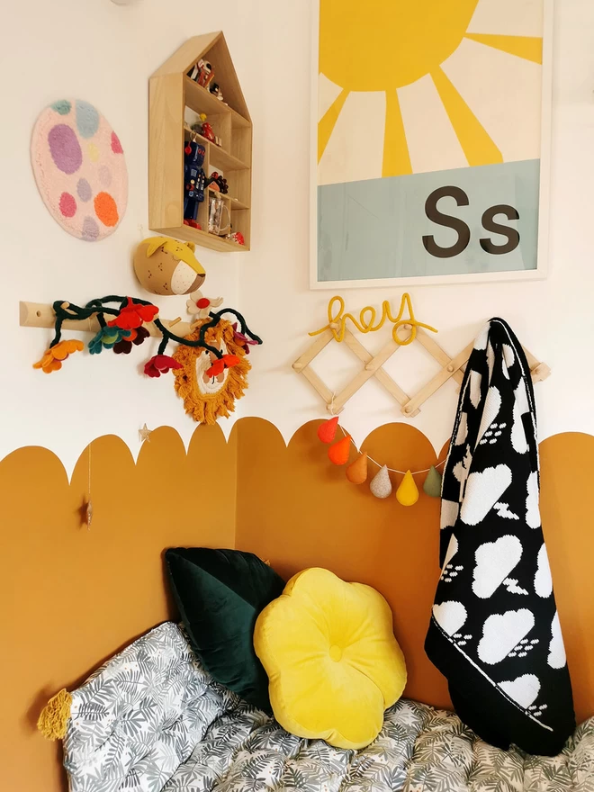 A modern childrens room with a cosy cushioned reading corner and half scalloped ochre walls. The walls are adorned with fun childrens decor, and on a wall peg hangs a black and white knitted storm cloud baby blanket.