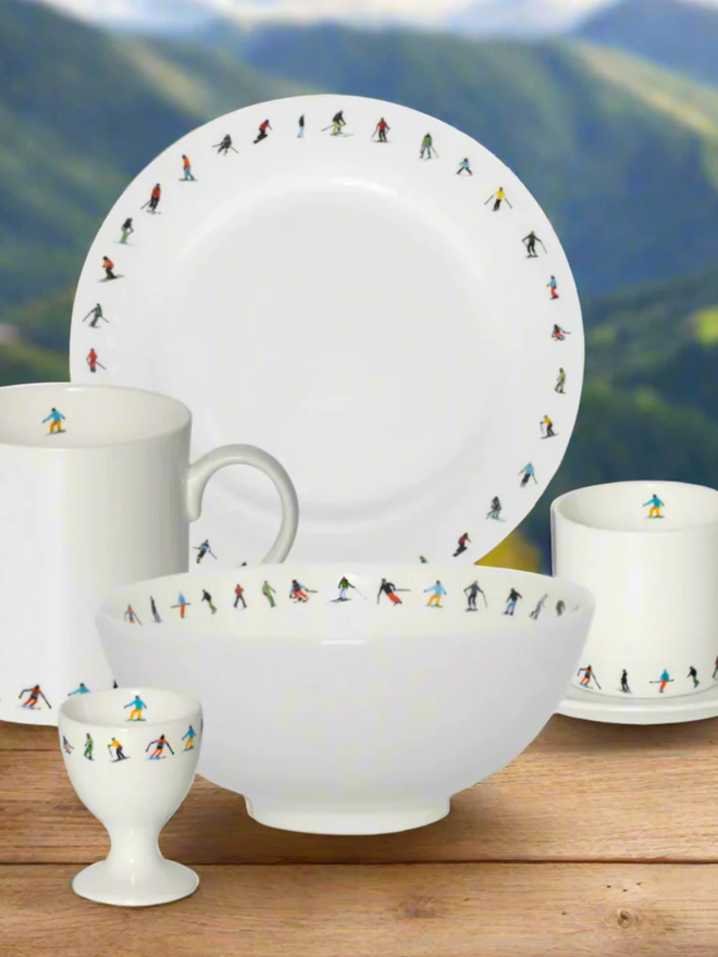 Ski Chain Breakfast Set. A side plate, a bowl, an egg cup, an espresso cup, and a saucer. All are a plain white with a pattern of small, multicoloured skiers. They are set on a wooden table against a green, mountainous background.