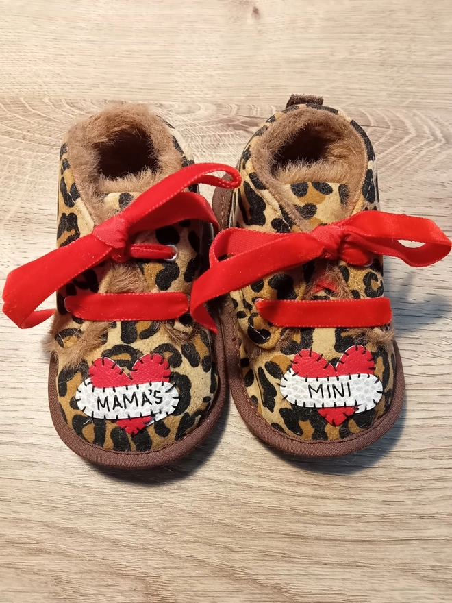 A pair of leopard print baby booties, lined with fur and with red velvet ribbon laces. A red heart with a white scroll adorns each booty. One says MAMA'S, the other MINI in black stitches