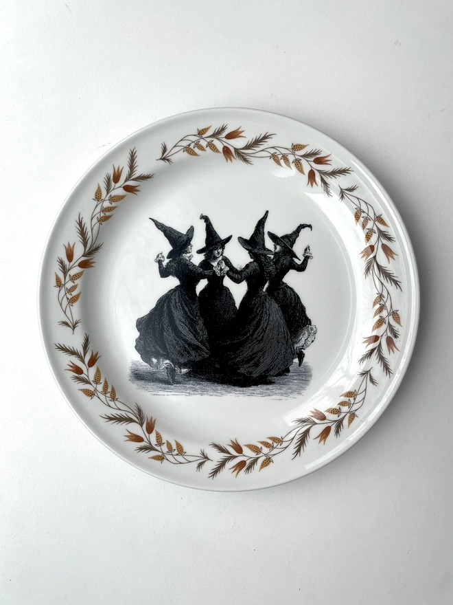 a coven of 4 witches dance hand in hand, the image is victorian in black and white and is printed directly onto an ornate vintage plate