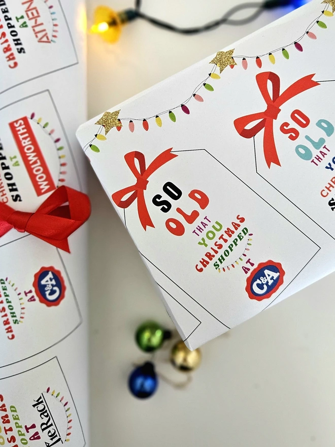 A photograph of a present wrapped in paper that has tags with a red bow printed on it. On each tag says so old that you Christmas shopped at Woolworths, Athena, C&A, Tie Rack, Sock Shop illustrated using brightly coloured text and the retro shop logo.