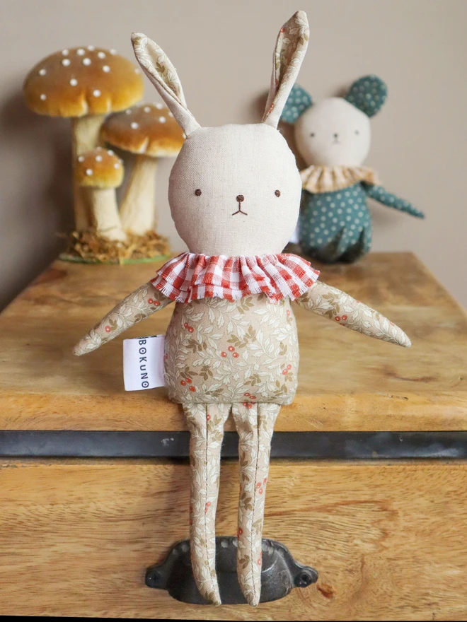 Handmade rabbit doll with light floral-patterned fabric, a red-and-white gingham ruffle collar. A white tag with the brand name ‘BOKUNO’ is sewn on the side.