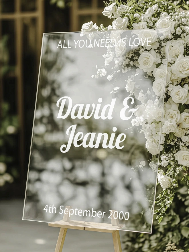 a clear pvc board leaning on a board holder with the wording all you need is love, David and Jeanie and the wedding date in white lettering, the board is standing infront of a bouquet of white flowers with green foliage