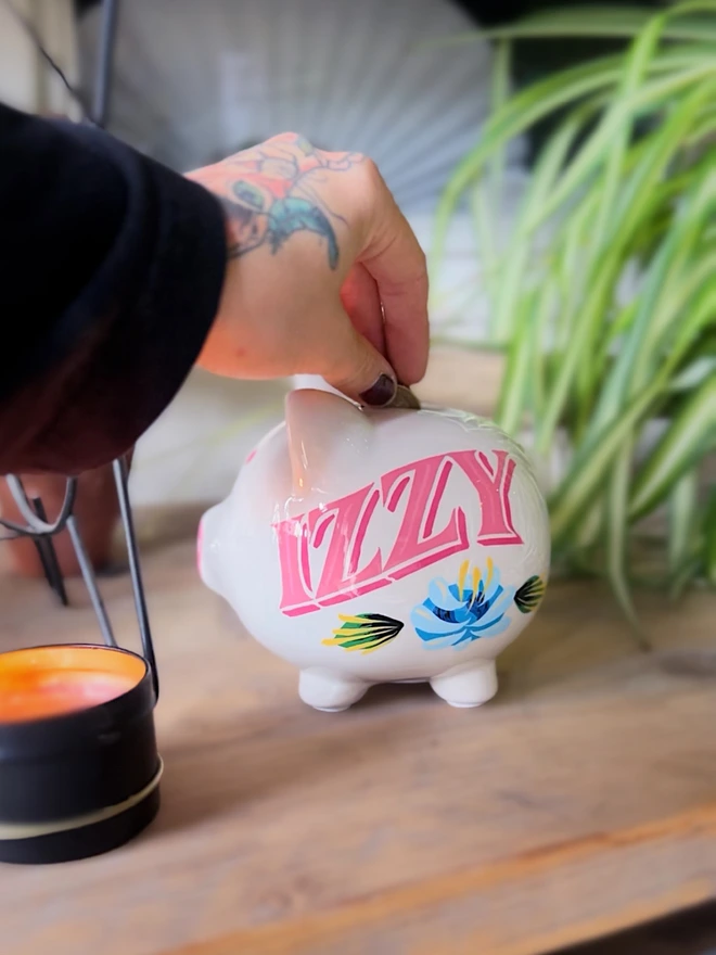 Hand-Painted piggy bank spelling 'Izzy' in pink lettering with a blue canal rose and green leaves.