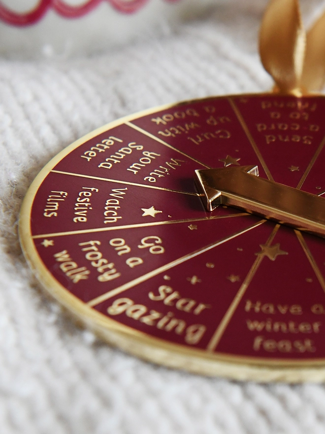 A deep red and gold Christmas decoration lays on a woollen blanket. It has 12 segments, each one with a different Christmas activity idea, and a golden arrow in the centre.