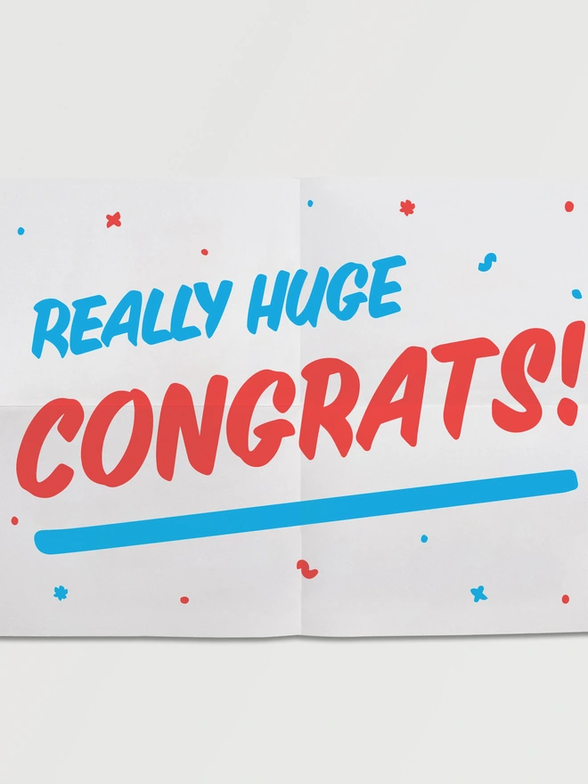 Unfolded A5 piece of paper that says REALLY HUGE CONGRATS! in red fun slanted typography on an angle with a blue underline