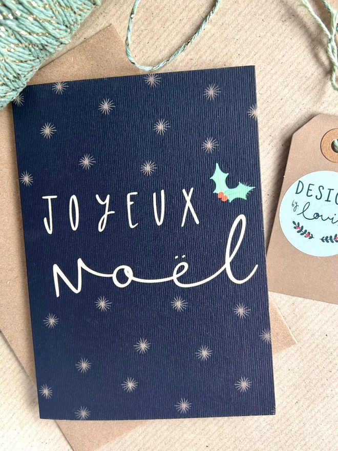 A dark navy background with white snowflake illustrations scattered across the card. In the middle reads the French 'Joyeux Noël' in white handwritten capital letters, with a small sprig of green and red holly to the side.