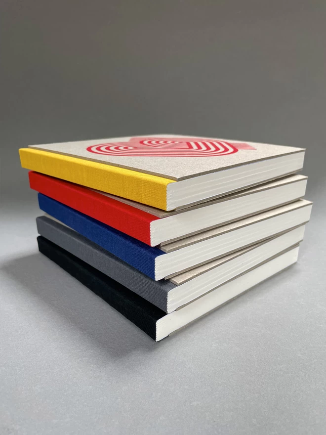A pile of pasteboard sketchbooks, colourful fabric spines towards us, white blank paper showing along their bottom edge. Set in a light grey studio shot.