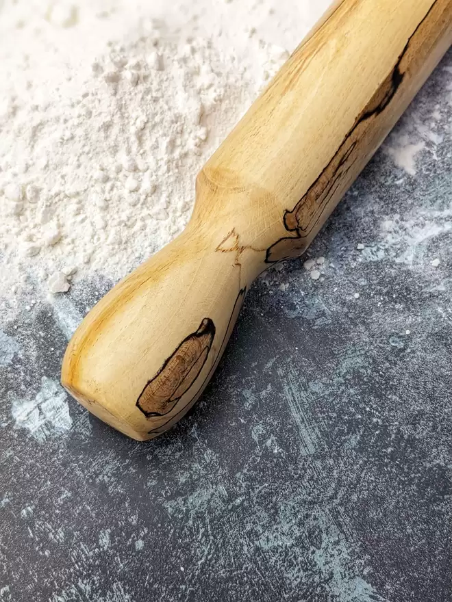 A highlight shot of a hand made rolling pin in Spalted Beech by Something From The Turnery, the picture is focussed on the handle of the rolling pin which boasts a unique darker brown grain pattern set on it’s more traditional golden Beech colour.