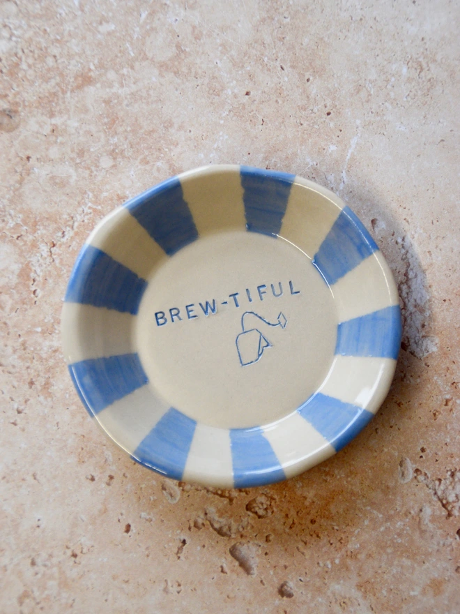 Handmade ceramic tea bag dish with blue striped rim and “Brew-tiful” text and teabag illustration in the centre, placed on a neutral stone background.