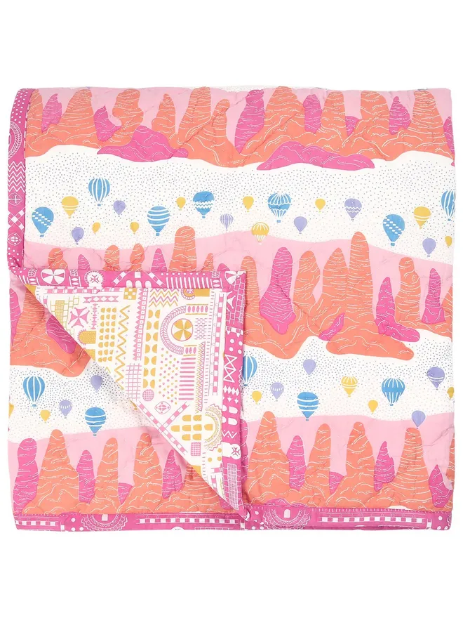 Fairy Chimney Quilt, a pink, orange and yellow quilted bedspread featuring a a colourful pattern of a landscape with hot air balloons in the sky, folded on a white background.