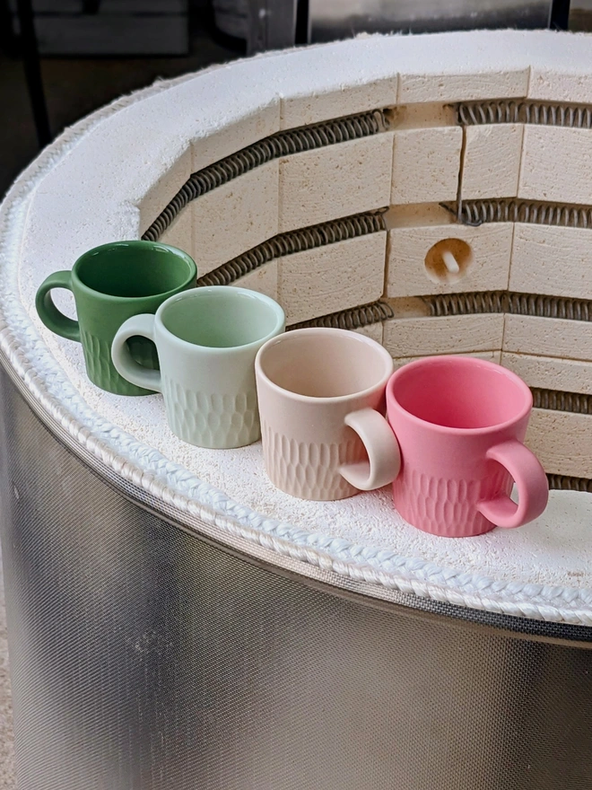 Four Tiny Espresso Cups in Dark Angelica Green, Pale Rockpool Green, Neutral Pale Oat and Milkshake Pink sat on the edge of a kiln in pottery studio