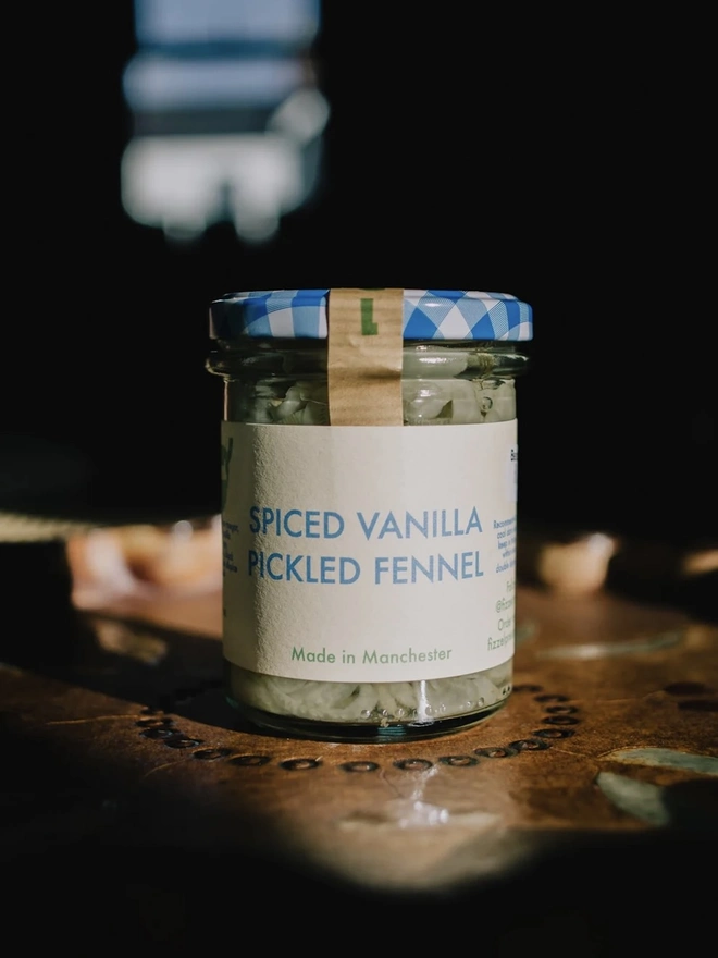 Jar Of Spiced Vanilla Pickled Fennel, jar of sauce with a blue lid on a decorated plate on a table. 