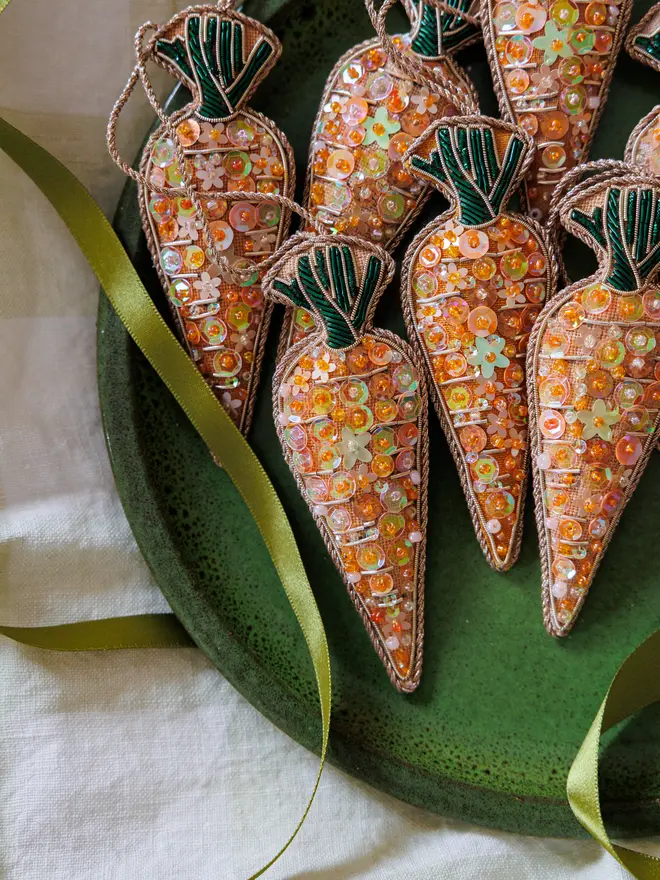 Multiple embroidered sequin carrot decorations laid on a dark green plate atop a white tablecloth, with green ribbon scattered around 