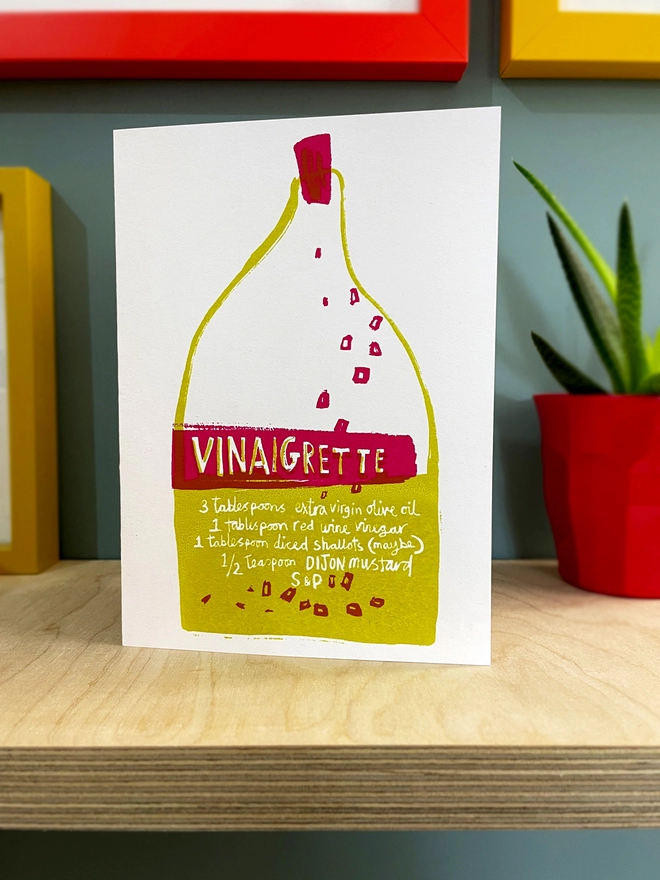 Vinaigrette bottle screenprinted with recipe drawn on the label, handprinted in green and red. Stands on a plywood shelf with coloured picture frames around and a plant to one side.