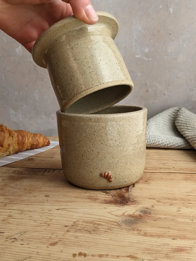 Croissant Butter Crock in biscuit glaze with a small croissant design on the front, set against a grey background.