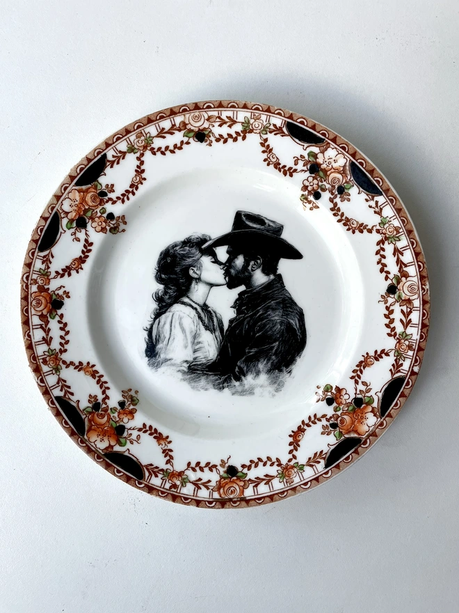 a vintage plate with an ornate border has a black and white vintage illustration of a cowboy and cowgirl sharing a kiss in the centre
