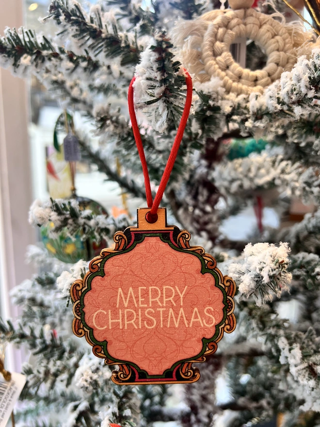 Multi coloured wooden tree decoration with 'Merry Christmas' written at the centre.  It is hanging by a red ribbon on a fake white christmas tree with other decorations. 