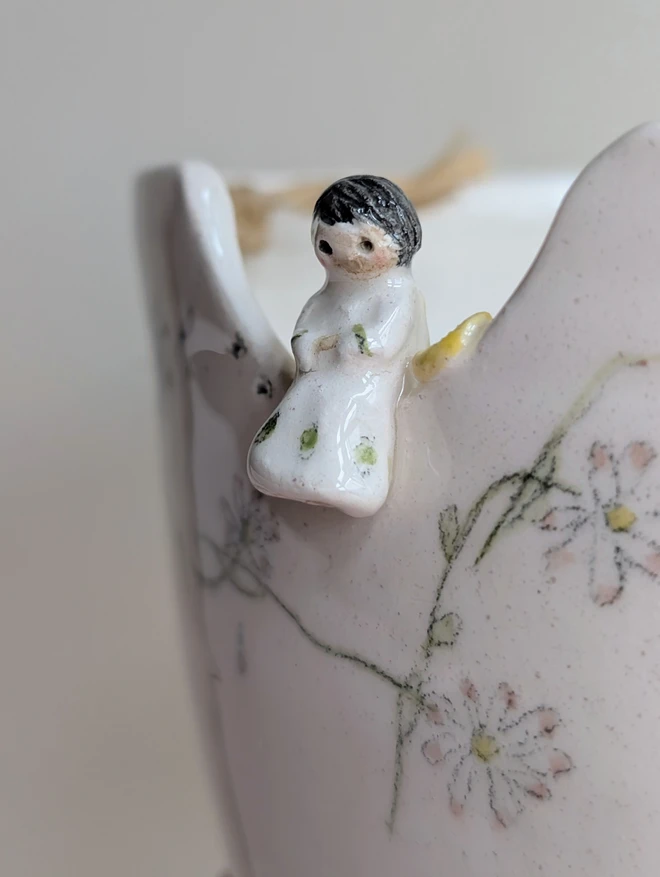 Close up of a tiny ceramic angel sitting on the edge of a planter with hand painted daisies on white clay