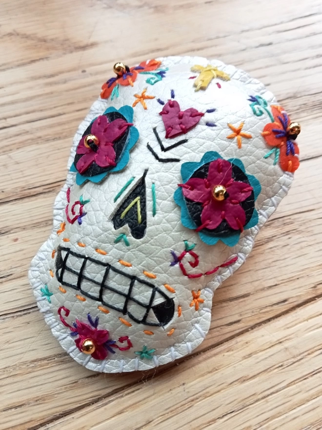 A hand stitched faux leather white Mexican Day of the Dead inspired sugar skull brooch with  colourful stitching and gold bead details, on a wooden background