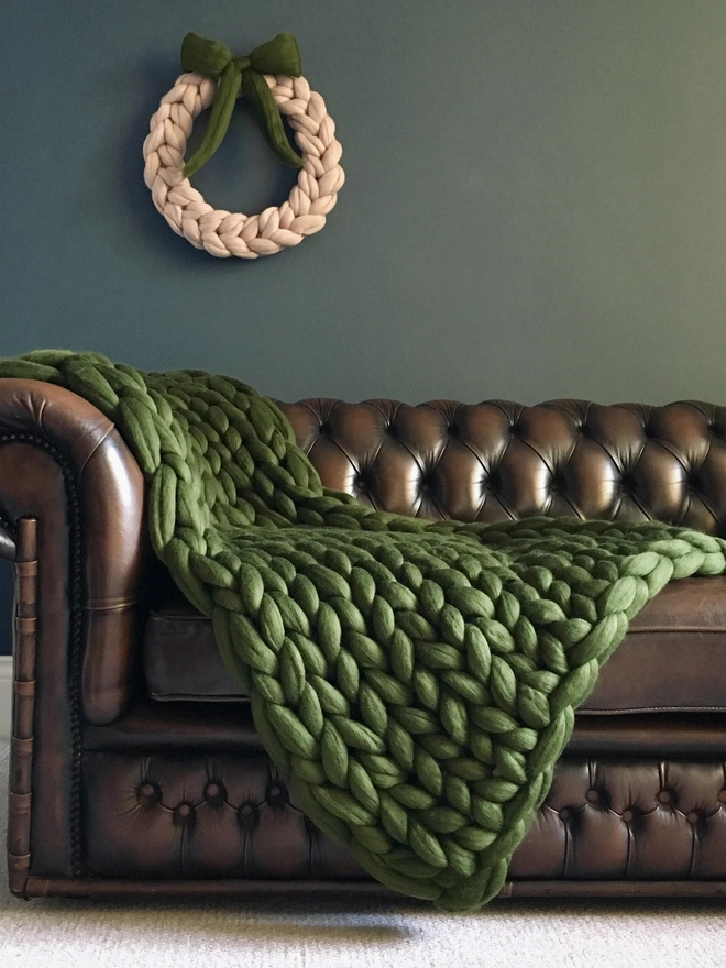 a willow green merino arm knitted blanket cascading down the side of a brown leather Chesterfield sofa, before a dark blue wall