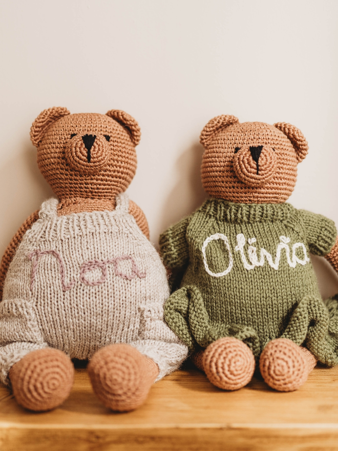 two hand crochet brown teddy bears on wooden shelf, wearing hand knit personalised beige dungarees and olive green dress