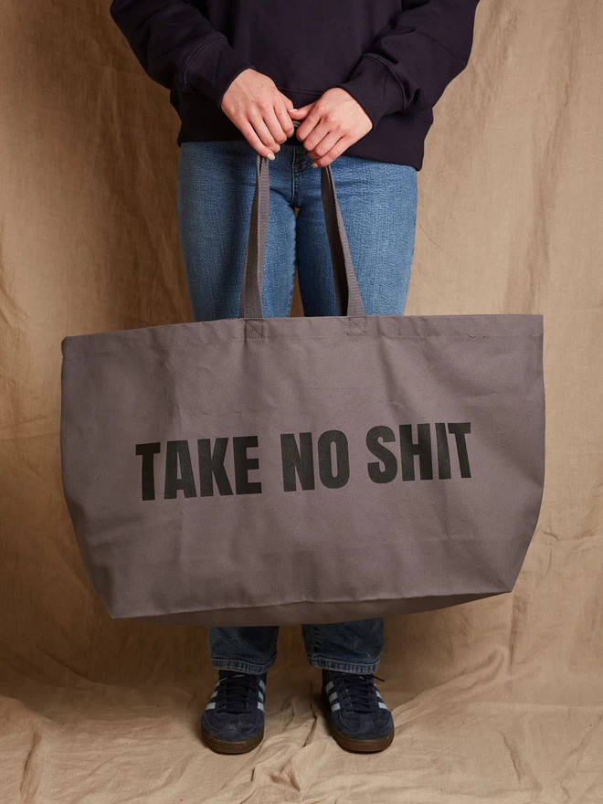 Model holding a massive gunmetal grey cotton canvas tote bag with the words Take No Shit printed in black 
