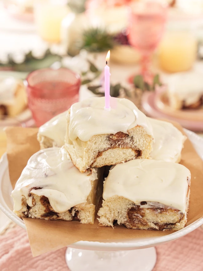 Birthday cinnamon buns on a cake stand with a candle in the top bun's vanilla frosting