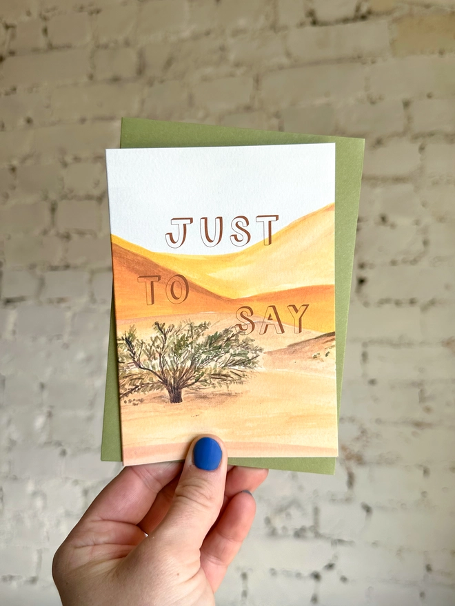 a greetings card featuring an illustration of a desert landscape with the text “just to say” with a green envelope
