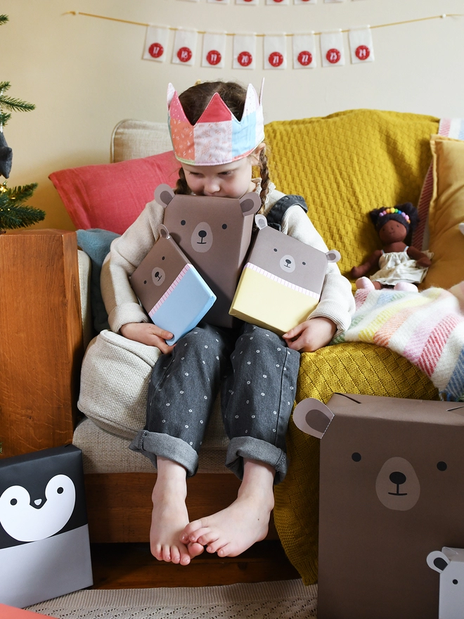 A young girl sitting on a sofa beside a Christmas Tree is holding three gifts wrapped up as brown bears.