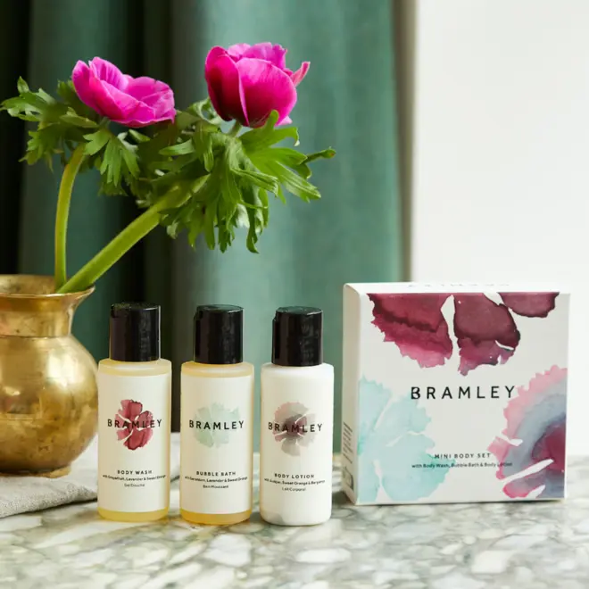 three small bottles of product, with white labels and black lids, next to a white box with blue and purple floral designs, and a gold vase with green-stemmed pink flowers, on a marble table with green curtain in background