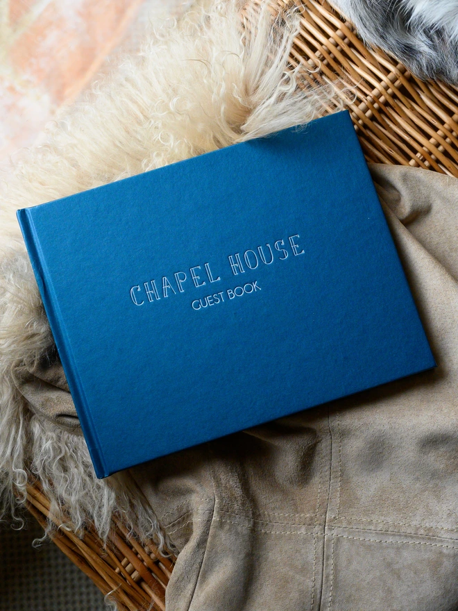 A navy Personalised Hardback Visitors Book with Outline Font rest on a table covered in a sheepskin.